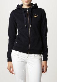 Sudadera con cremallera color azul marino, hecha de una mezcla de algodón, con una capucha interior a rayas, un logo dorado en el pecho y detalles con cordón. Textura suave.