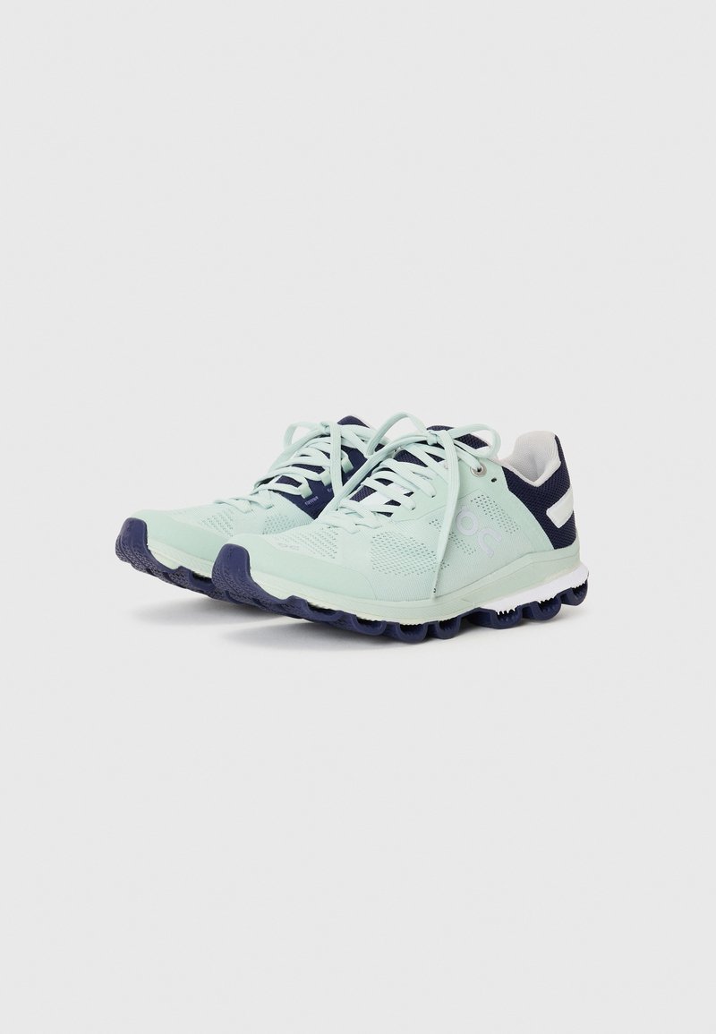 Hellgrüne Laufschuhe mit einem strukturierten Mesh-Obermaterial, marineblauen Akzenten und einer gepolsterten Sohle mit ausgeprägten Gummigriffen. Die Schnürsenkel sind intakt.