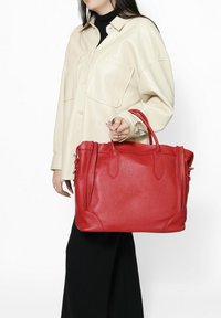 Borsa tote in pelle rossa con superficie testurizzata, doppi manici superiori e forma strutturata. Indossata con una camicia beige a bottoni e pantaloni neri.