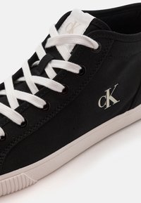 Svart canvas sneakers med vita snören, med en gummisula och en krämfärgad logotyp 'cK' på sidan. Synliga kontrastsömmar.