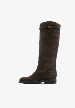 Botte haute en cuir suédé marron foncé avec un petit talon et un détail de boucle dorée près du haut, vue de côté sur fond blanc.