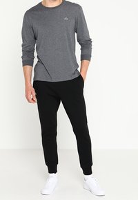 Camisa de manga larga gris con un pequeño logotipo verde, combinada con pantalones de chándal negros ajustados y zapatillas blancas sobre un fondo neutro.