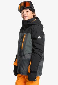 Veste de ski noire et grise avec des accents, des fermetures éclair et une capuche orange. Associée à un pantalon de neige orange et des gants noirs. Lunettes de ski sur la tête.