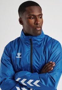 Mann mit verschränkten Armen, der eine blaue Hummel-Sportjacke mit Reißverschluss und weißen Chevronstreifen an den Ärmeln trägt, blickt zur Seite.