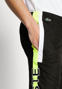 Svarte treningsbukser med hvite og neongule sidestriper; har en lomme og et logoakzent. Laget av lett stoff.
