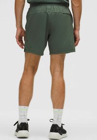 Uomo che indossa pantaloni sportivi verde scuro, calzini bianchi Lululemon e scarpe nere, visto di spalle su uno sfondo semplice.