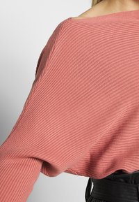 Maglione oversize a costine in rosa polveroso, con ampie maniche e scollo a barca. Il tessuto strutturato conferisce profondità al design.