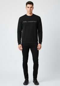 Sudadera negra con "KARL LAGERFELD" impreso en gris, cuello redondo y mangas largas. Combinada con pantalones ajustados negros y zapatos negros.