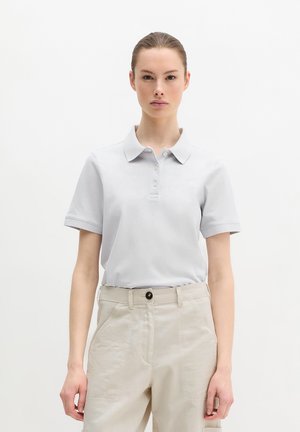 Junge Frau mit zurückgebundenem Haar, die ein hellgraues Poloshirt mit kurzen Ärmeln trägt, das in beige hochtaillierte Hosen gesteckt ist, vor einem schlichten Hintergrund.