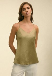 Olijfgroene satijnen cami met een V-hals, dunne bandjes en een gladde textuur, gecombineerd met lichtgekleurde broek.