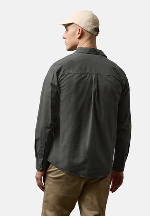 Mann trägt ein dunkelgraues Langarmshirt, beige Hose und eine beige Baseballmütze, steht mit dem Rücken zu einem schlichten hellen Hintergrund.