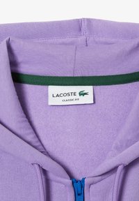 Felpa con zip viola con colletto interno verde. Etichettata "LACOSTE CLASSIC FIT." Tessuto morbido con una trama liscia.