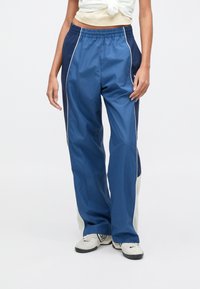 Μπλε σκούρο παντελόνι φόρμας με ελαφρύ, λείο ύφασμα. Διαθέτει λευκή περιμετρική ρίγα και αντίθετο ανοιχτοπράσινο πάνελ στο κάτω μέρος του ποδιού. Ελαστική μέση.