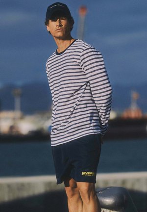 Pasiasta koszula z długim rękawem w białym i granatowym kolorze, zestawiona z granatowymi szortami z żółtym napisem. Model nosi granatową czapkę na tle nadmorskim.