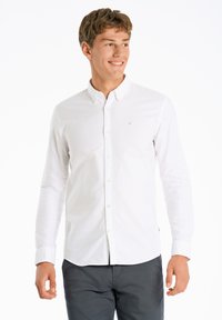 Witte blouse met lange mouwen, voorzien van een kraag en een klein embleem op de borst. Gemaakt met donkergrijze broek. Gladde textuur.