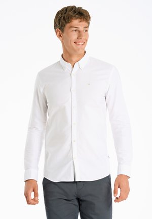 Witte blouse met lange mouwen, voorzien van een kraag en een klein embleem op de borst. Gemaakt met donkergrijze broek. Gladde textuur.