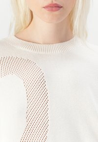 Ljusbeige stickad tröja med ett stort öppet stickmönster på ena sidan. Rundhalsen har en ribbad textur och själva tyget är slätt.