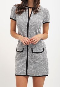 Šedé šaty v herringbonovém vzoru s krátkými rukávy, černými lemy, výstřihem ve tvaru klíčové dírky a dvěma předními kapsami s kovovými prvky ve stříbrné barvě.