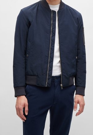Homme portant une veste bomber bleu marine sur une chemise blanche et un pantalon bleu marine, debout avec les bras détendus devant un fond gris.