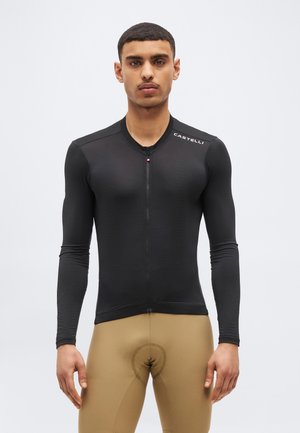 Moški v oprijetem črnem kolesarskem dresu z dolgimi rokavi z logotipom "Castelli" in rjavimi oblazinjenimi kolesarskimi hlačami stoji pred enobarvnim ozadjem.