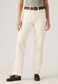 Άτομο που φοράει εκρού παντελόνι με ίσια γραμμή, καφέ δερμάτινη ζώνη, γκρι μπλούζα και σκούρα καφέ δερμάτινα παπούτσια με κορδόνια, στέκεται μπροστά σε απλό φόντο.