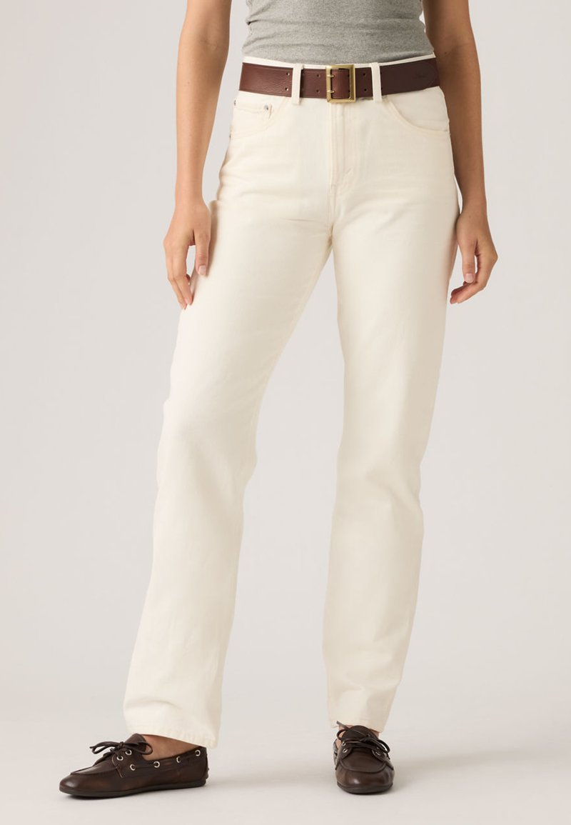 Άτομο που φοράει εκρού παντελόνι με ίσια γραμμή, καφέ δερμάτινη ζώνη, γκρι μπλούζα και σκούρα καφέ δερμάτινα παπούτσια με κορδόνια, στέκεται μπροστά σε απλό φόντο.