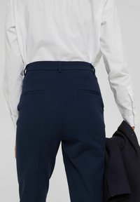 Personne portant un pantalon bleu foncé sur mesure et une chemise blanche à manches longues, tenant un blazer foncé dans sa main droite.