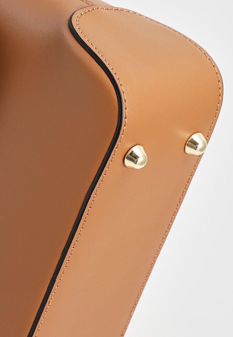 Taschenhandtasche aus tanfarbenem Leder mit abgerundeten Kanten, schwarzem Rand und glänzenden goldenen Hardware-Akzenten. Strukturierte Oberfläche mit sichtbaren Steppnähten.