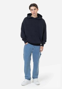 Sweatshirt azul-marinho com capuz ajustável, bolso frontal e punhos canelados, combinado com jeans azul claro e ténis brancos. Design simples.