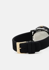 Correa de reloj de goma negra con un acabado texturizado, que presenta una hebilla de metal dorado y una caja plateada en el lado opuesto.