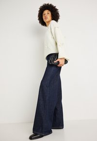 Kremo spalvos megztinis su kaspino detalėmis, derinamas su tamsiai mėlynomis plačiomis kelnėmis. Modelis laiko juodą tekstūruotą rankinę. Paprastas, modernus įvaizdis.