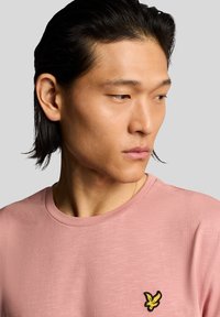 Lyserød t-shirt med rund hals, med et sort og gult broderet logo på venstre bryst. Blødt, tekstureret stof.