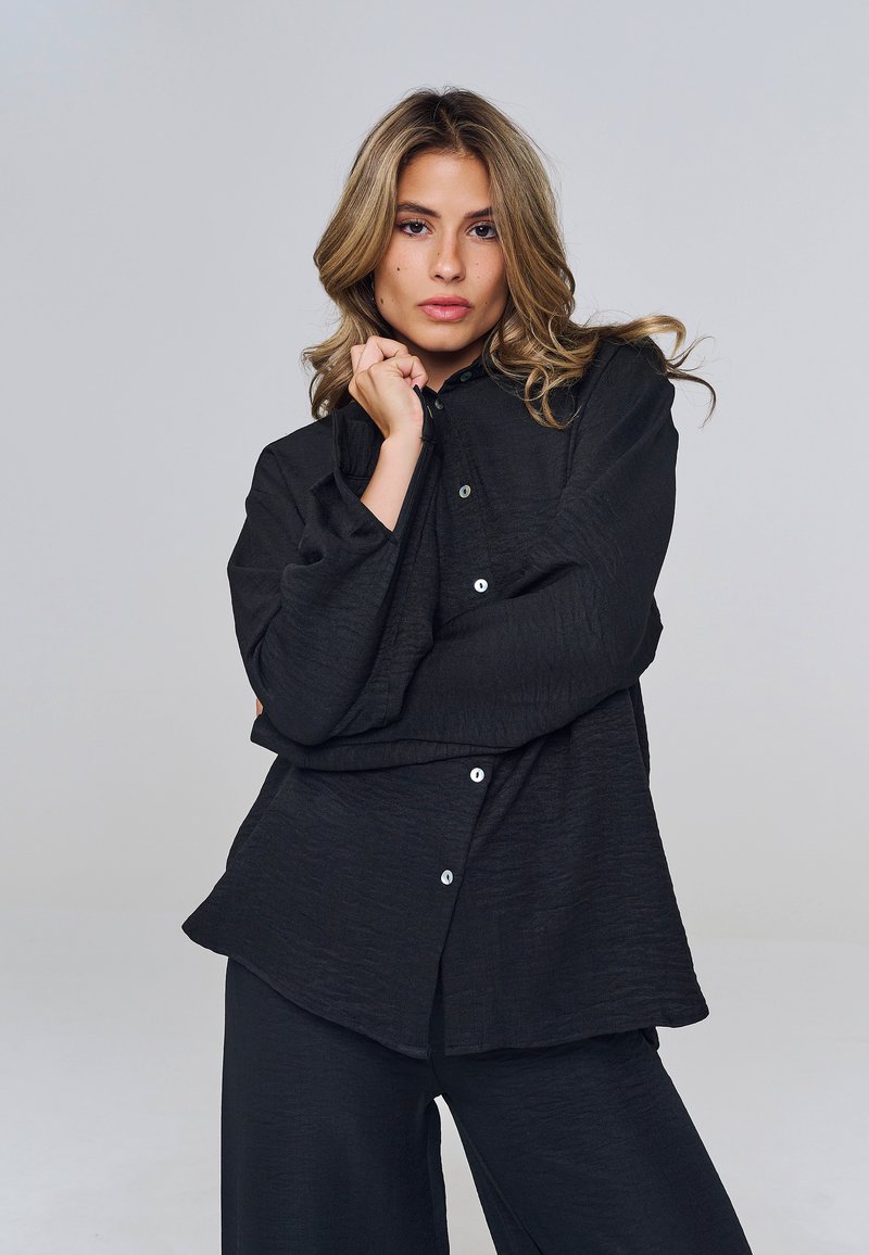 Femme aux cheveux blonds ondulés portant une chemise noire ample à boutons et un pantalon large assorti, posant avec une main près du visage.
