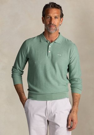 Lichtgroene gebreide polo trui met een geribbelde structuur, voorzien van een kraag en een drieknoopssluiting. Gecombineerd met lichte broek.