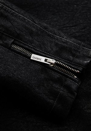 Chaqueta de mezclilla negra con superficie texturizada, que presenta una cremallera de metal con una etiqueta "OMBRE" y detalles de costura en contraste.