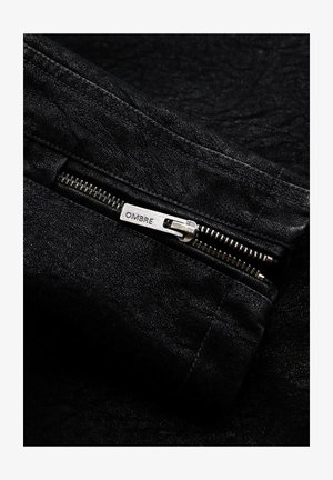 Schwarze Jeansjacke mit strukturierter Oberfläche, ausgestattet mit einem Metallreißverschluss mit einem "OMBRE"-Label und kontrastierenden Stickdetails.