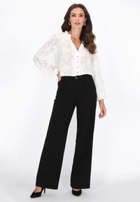 Blusa blanca de manga larga con cuello con volantes y patrón sutil, combinada con pantalones negros de cintura alta y tacones negros.