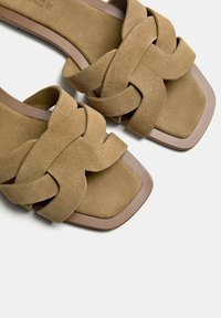 Taupe suede slide sandals with a woven strap design and a square toe box. Features a smooth brown sole and minimalistic aesthetic.