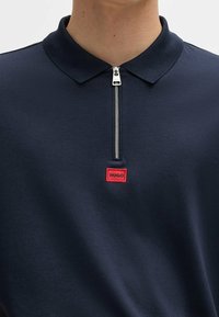 Námořnická polo košile vyrobená z měkkého materiálu s límcem na zip. Na hrudi se nachází červená obdélníková nášivka s nápisem "HUGO".