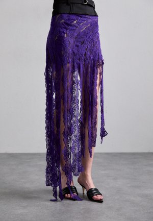 Lila Spitzenrock mit fransigen Kanten, der aufwendige Blumenmuster zeigt. Getragen mit schwarzen, offenen, hohen Sandalen. Der Hintergrund ist grau.