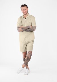 Man met tatoeages op zijn armen en benen staat met zijn armen gekruist, gekleed in een beige shirt met korte mouwen, bijpassende korte broek en witte sneakers tegen een witte achtergrond.