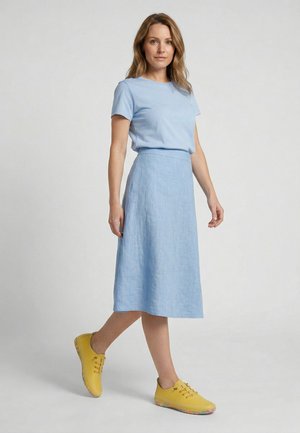 Woman standing wearing a light blue short-sleeve top and matching midi skirt with bright yellow lace-up shoes.