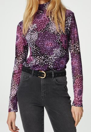 Frau trägt ein lila, langärmliges, blumengemustertes Netzoberteil, das in eine hoch taillierte schwarze Jeans mit einem schwarzen Gürtel mit goldener Schnalle gesteckt ist.