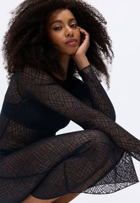 Vestido preto transparente com padrão geométrico, mangas longas e silhueta ajustada. Modelo posa com a mão no queixo, destacando a textura da peça.