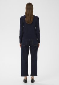 Sötétkék, slim fit hosszú ujjú pulóver, magas derekú, bőszárú sötét farmernadrággal és fekete lapos cipővel, hátulnézetből.