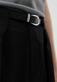 Zwarte stoffen broek met een wijde pijp, voorzien van een zwarte leren riem met een zilveren gesp. De riem heeft gaten voor verstelbaarheid.