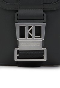 Fekete bőr kézitáska ezüst fém csattal, amelyen a "KL" kezdőbetűk találhatók. A pánton egy texturált szalag díszítés található.