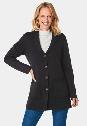 Langer schwarzer Cardigan aus strukturiertem Strickstoff, mit Schalkragen, vorderen Knöpfen und zwei aufgesetzten Taschen, getragen über einem hellen Rollkragenpullover.