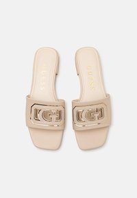 Bežové slip-on sandále so štvorcovou špičkou, lesklým zlatým logom a hladkou syntetickou textúrou. Názov značky je vyrazený zlatom na stielke.