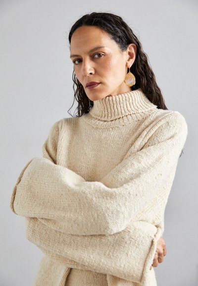 Μπεζ χοντρή πλεκτή μπλούζα με υψηλό ριμπ στο λαιμό και oversized μανίκια. Λείο υφασμάτινο σχέδιο, ελάχιστες λεπτομέρειες.